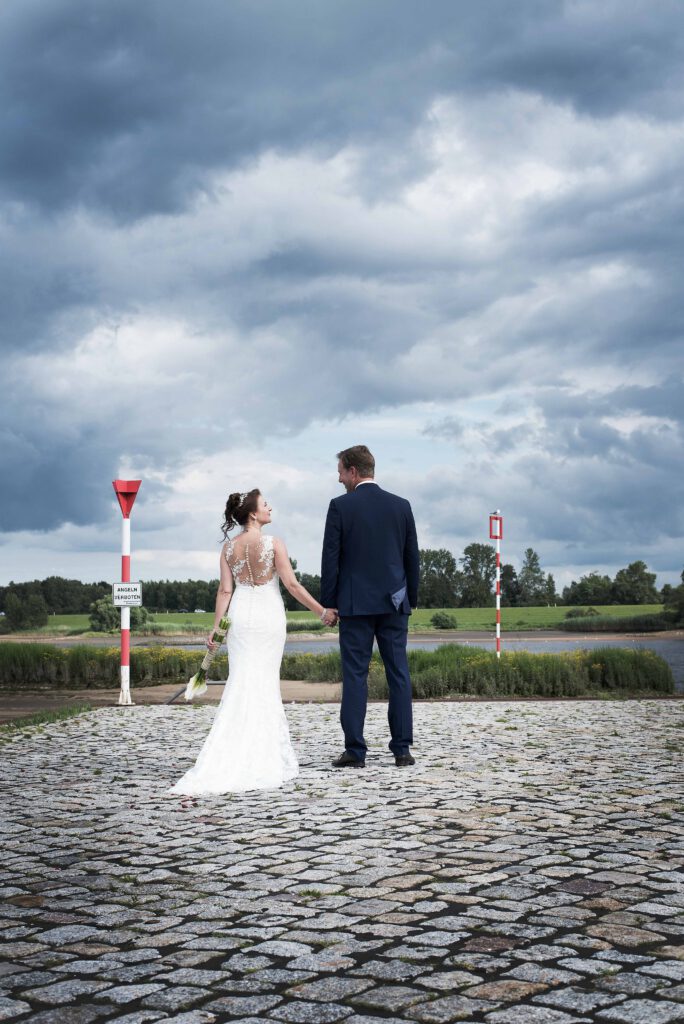 Brautpaar von Hinten, Hand in Hand, vor bewölktem Himmel am Wasser, von Stark wie Zwei Hochzeitsfotografie